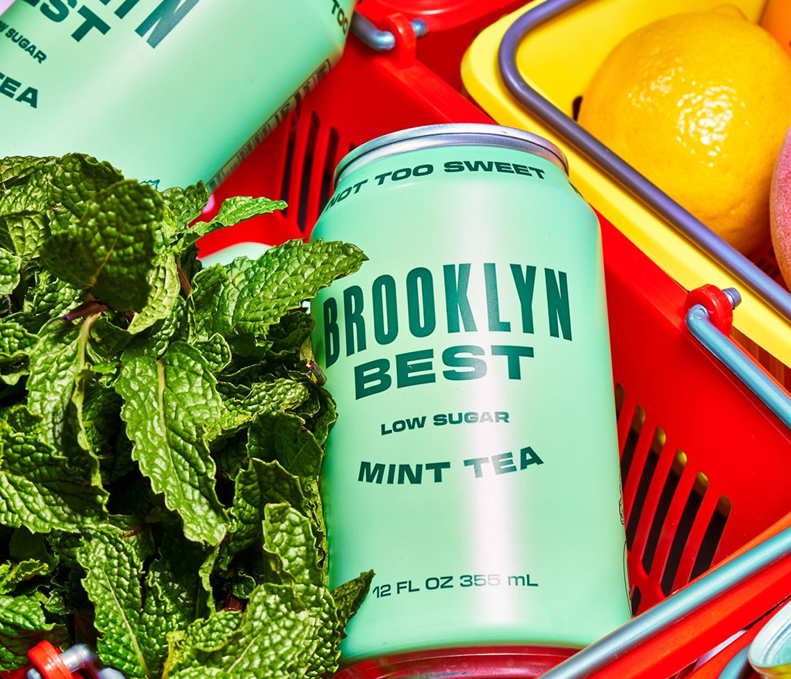 Brooklyn Best Mint Tea can in a red basket, next to fresh mint leaves and lemons. Green can reads Low Sugar, Not Too Sweet.