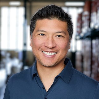 Smiling man with short black hair in a dark blue collared shirt stands in a bright room with shelves and large windows behind him.