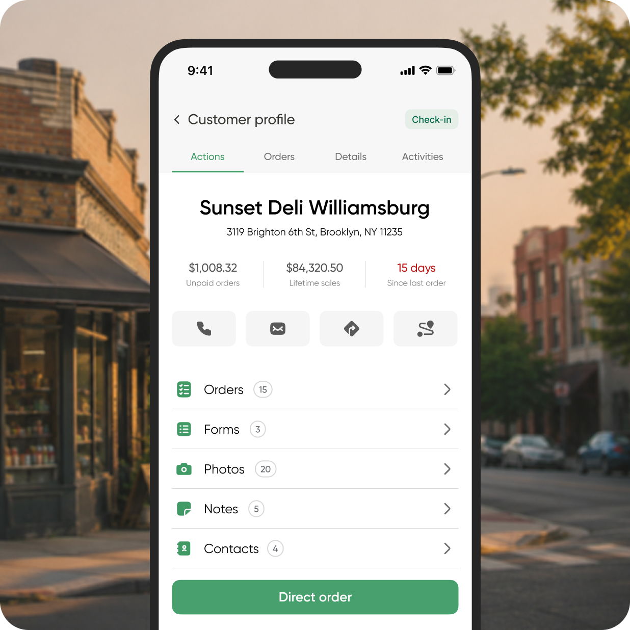 A smartphone held up on a bustling city street lined with shops and trees shows the V3 Sunset Deli Williamsburg customer profile on its screen, featuring finance details, sales data, order history, and contact information prominently on the front page.