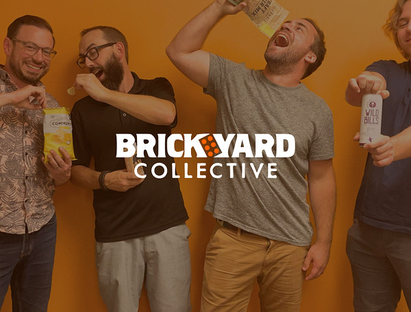 Four men are gathered in front of a vibrant orange wall, sharing laughter and snacks as they each hold a different package of food. Their relaxed, joyful expressions reflect genuine camaraderie, embodying the essence of a memorable Front Page moment. Bold white text overlay reads “BRICKYARD COLLECTIVE,” highlighting the energetic and welcoming atmosphere of this V3 group gathering.